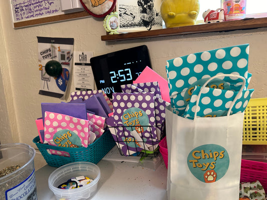A collection of paper bags, pink, purple and blue. Some have polka dots on them. In the front is a larger paper bag holding the blue bags with polka dots. All bags have a sticker on them that says "Chips Toys" with a drawing of an orange paw print below. in the background is a digital clock and other random stuff.