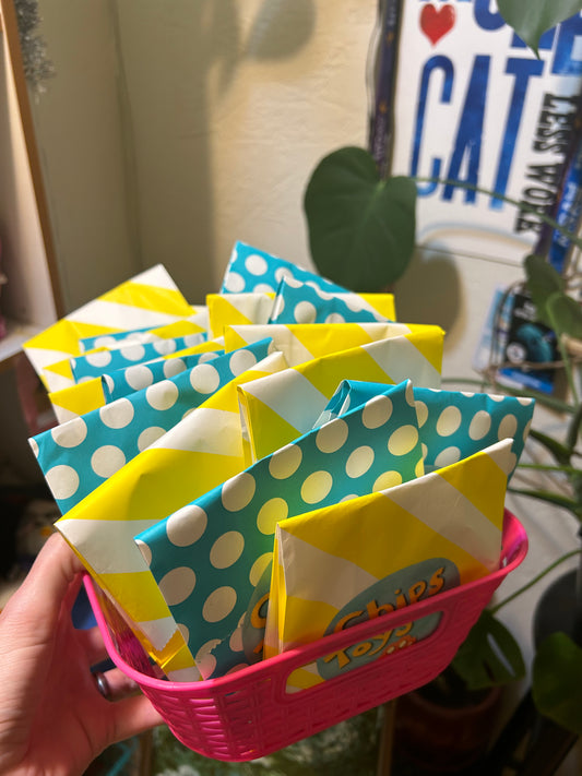 A person holding a pink basket of paper bags. The bags are blue with polka dots and yellow with stripes. There are stickers on the bags that say "Chips Toys". In the background are some houseplants and a sign with a red heart and the word "Cat" on it.