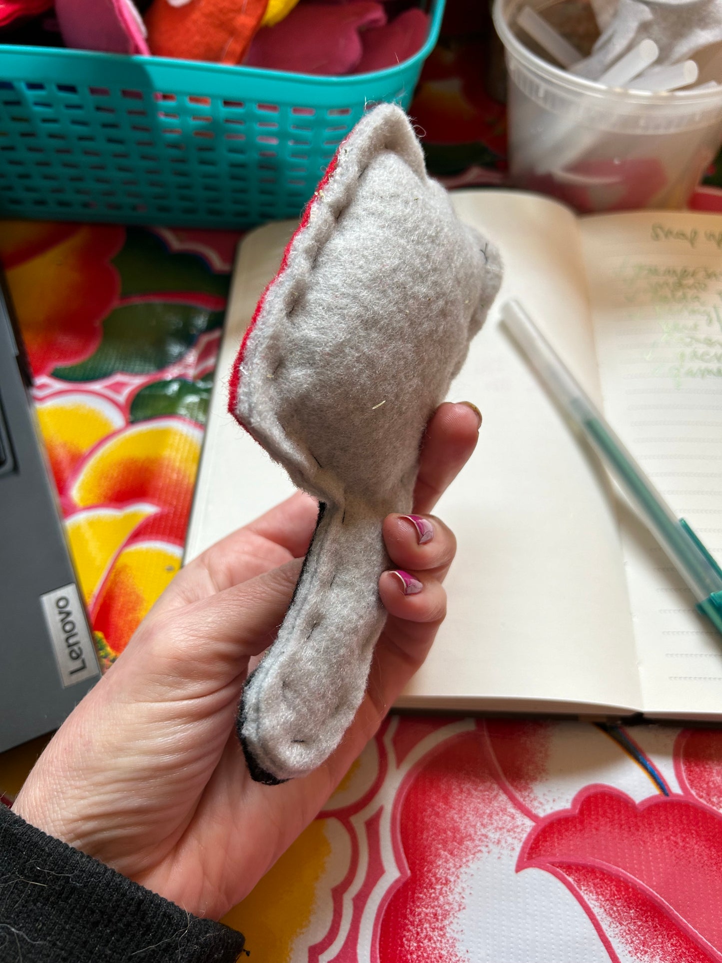 A person holding a catnip cat toy for sale. The view of it is the side/back, it is grey felt, cut in the shape of a cleaver. There is a notebook and pen in the background.