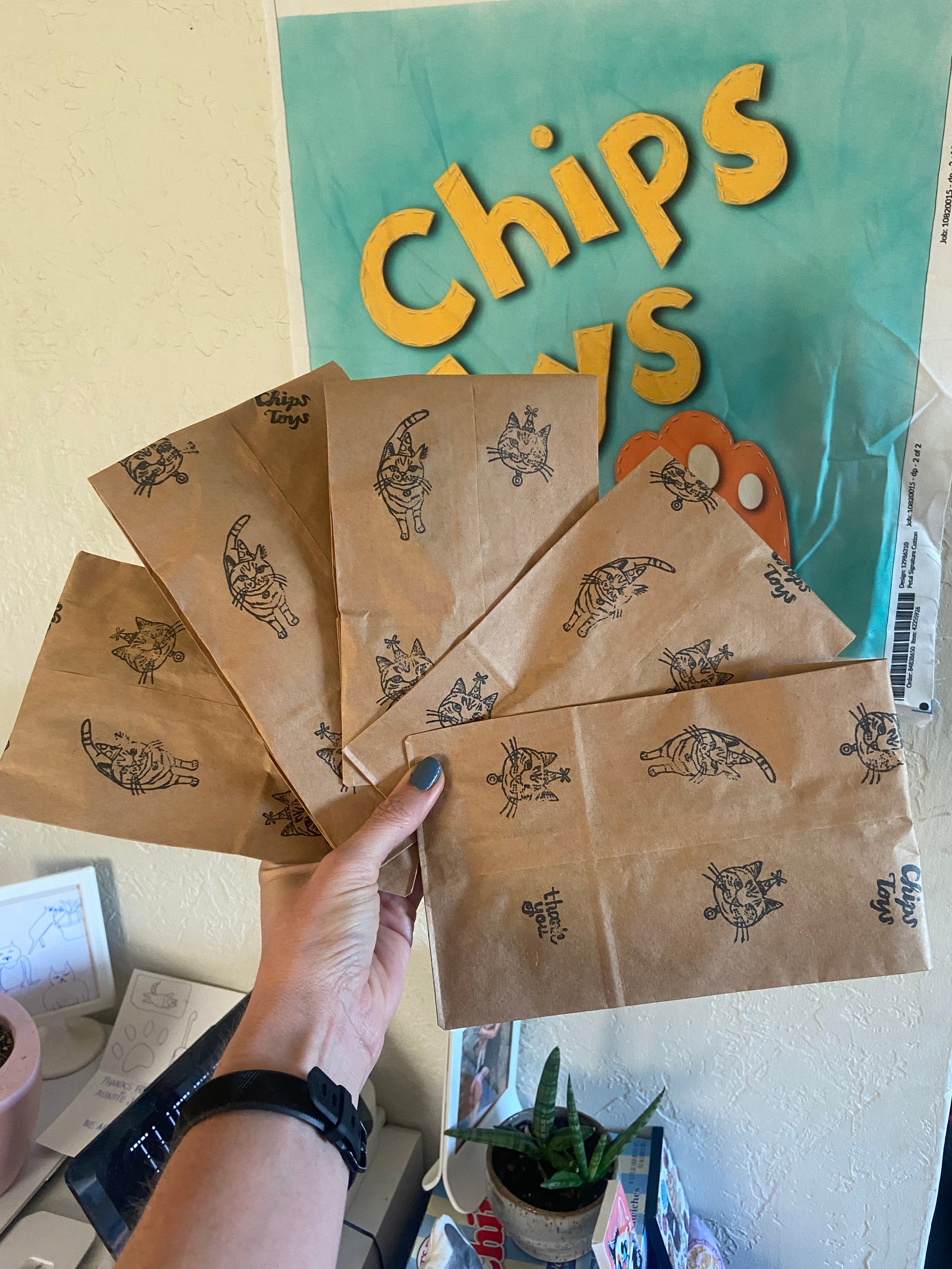 A person holding five brown paper bags with stamps of cats on them. In the background there is a blue banner on the wall that says "Chips Toys".