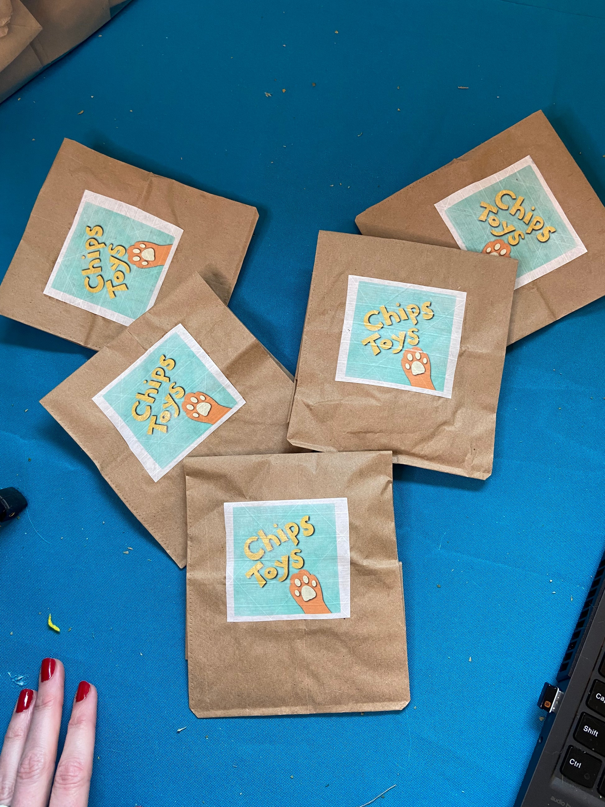 A group of 5 brown paper bags on a blue tablecloth. On each bag is a square sticker that says "Chips Toys" with a drawing of an orange cat paw below that.