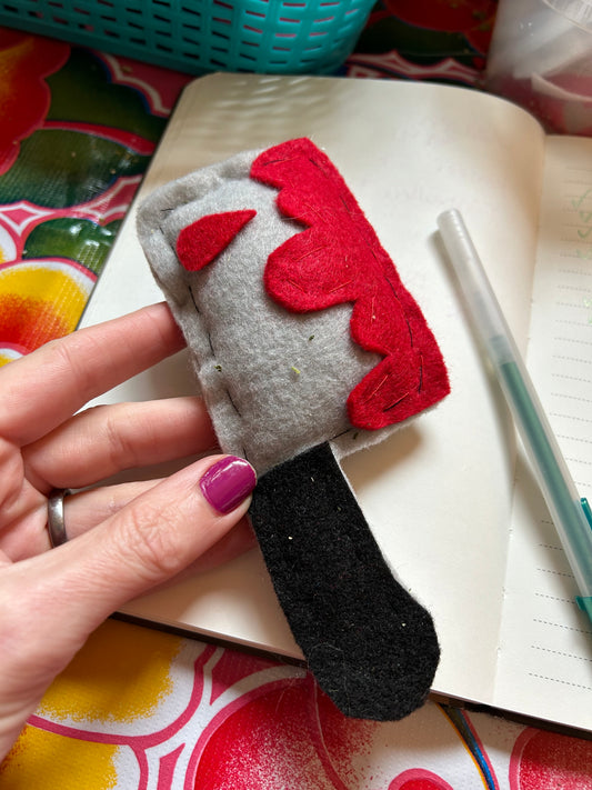 A person holding a catnip cat toy for sale. The toy is made of felt and is in the shape of a cleaver with blood on it. A notebook and a pen are in the background.