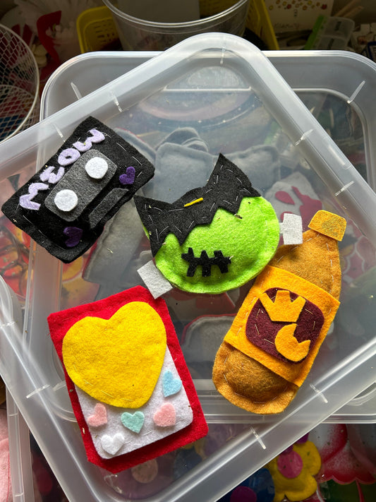 An assortment of felt catnip cat toys for sale. They are in the shape of a cassette tape that says "meow" on the top, a green frankenstiein head, a valentines candy box and a 40 oz of beer.